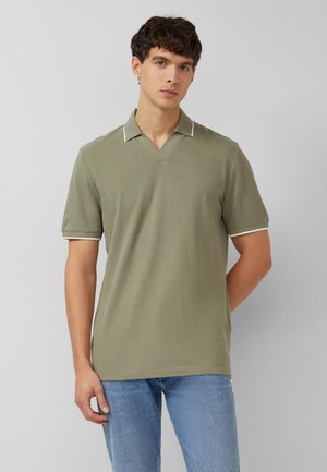 Jeune homme portant un polo à manches courtes de couleur vert olive clair et un jean bleu clair, debout devant un fond uni gris clair.
