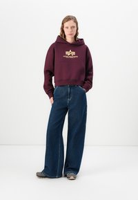 Felpa bordeaux con logo dorato "Alpha Industries", abbinata a jeans scuri a gamba larga e sneakers grigie. Design dell'outfit semplice e informale.