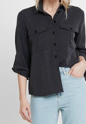 Femme portant une chemise noire à boutons avec les manches retroussées et un jean bleu clair, main dans la poche, sur un fond uni.