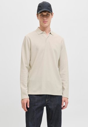Unge mand iført en beige langærmet polotrøje, mørke jeans og en sort kasket, står mod en ensfarvet lys baggrund.