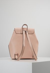 Kuusikulmainen posliininvaaleanpunainen nahkareppu, jossa säädettävät olkahihnat, sileä pinta ja minimalistinen muotoilu; varustettu läpällä ja hopeisilla metalliosilla.