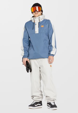 Chaqueta con capucha azul y blanca con cierre a presión en el frente, puños elásticos, combinada con pantalones blancos holgados y guantes negros. Zapatos negros con detalles blancos.