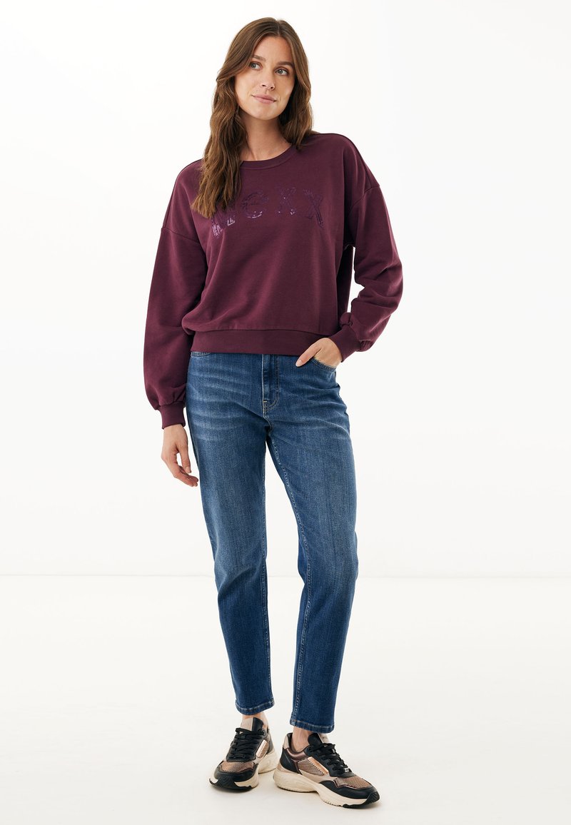 Bordeaux-coloured sweatshirt met ronde hals met grafische tekst, gecombineerd met blauwe rechtlopende jeans en zwart-witte sportschoenen. Soepele textuur.