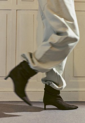 Persoană purtând pantaloni deschiși la culoare și ghete negre cu toc pentru glezne, mergând pe podea cu mochetă în interior.