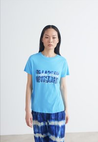 Light blue cotton t-shirt with a graphic print. Model wears tie-dye blue pants. Simple crew neckline and short sleeves.