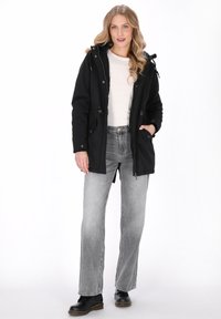 Kvinde med langt blond hår iført en sort hættejakke, hvid skjorte, bleget grå jeans og sorte støvler, stående foran en hvid baggrund.