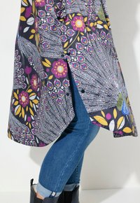 Tunique multicolore à motifs floraux et plumes de paon, fabriquée en tissu léger, associée à un jean bleu et des bottes noires.