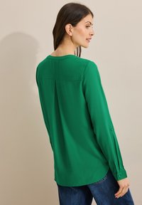 Blusa verde de manga larga hecha de tela suave, con un detalle de pliegue en la parte posterior y puños abotonados. Llevada con unos vaqueros de mezclilla azul oscuro.