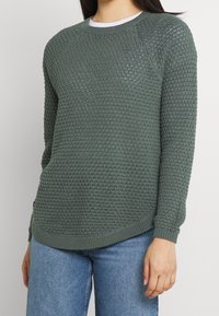 Grüner Strickpullover mit einem strukturierten, gewebten Muster. Verfügt über Raglanärmel sowie Rippbündchen und Saum, kombiniert mit hellblauen Jeans.