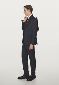 Mörkgrå skräddarsydd kostym med två-knappars kavaj och byxor med platt front. Bärs med vit skjorta, grå slips och svarta läderskor. Ryggsäck hängd över axeln.