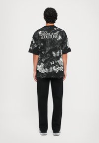 Čierna krátkoprstová košeľa s kvetinovými a motýľovými vzormi, na zadnej strane s textom "Versace Jeans Couture", v kombinácii s čiernymi nohavicami.