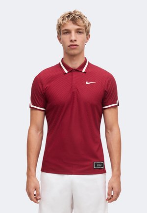 Jeune homme aux cheveux blonds courts et bouclés, portant un polo rouge Nike avec des bordures blanches et un short blanc, debout devant un fond uni.