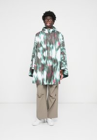 Patterned poncho in shades of teal, white, and pink, featuring a hood and wide sleeves. Paired with beige wide-leg pants and white shoes.