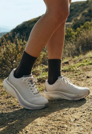 Jambes portant des chaussettes noires et des chaussures de course blanches, debout sur un sentier de terre avec une végétation sèche et des collines en arrière-plan.