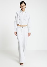 Frau trägt ein weißes, kurzes Hemd mit Knöpfen und Schleifendetails sowie eine hoch taillierte, weiße Hose mit weitem Bein und steht auf einem weißen Boden.