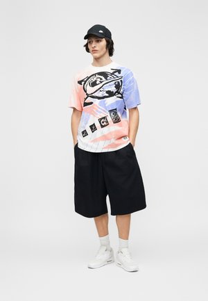 Jeune homme portant un t-shirt graphique multicolore, un short large noir, des baskets blanches, des chaussettes blanches et une casquette noire, debout les mains dans les poches.