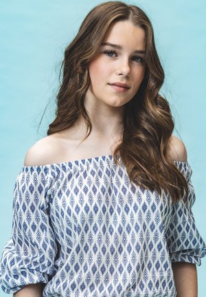 Witte off-shoulder blouse met blauw diamantpatroon. Zachte stof, pofmouwen en een ontspannen pasvorm. Neutraal blauwe achtergrond.