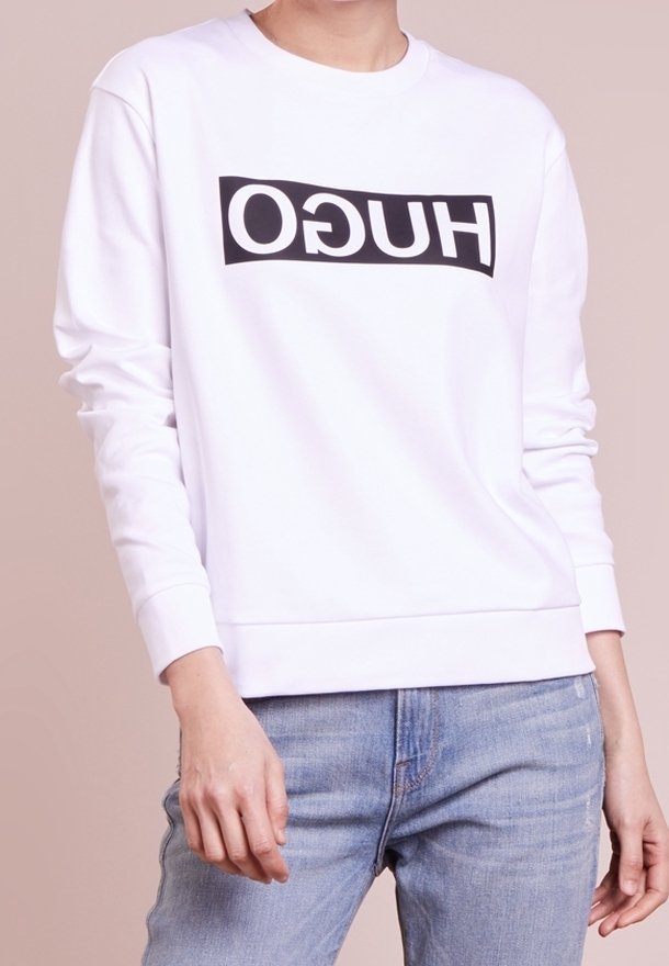 Osoba v bílém dlouhém rukávu s obráceným černým logem "HUGO" a světle modrými džíny na hladkém béžovém pozadí.