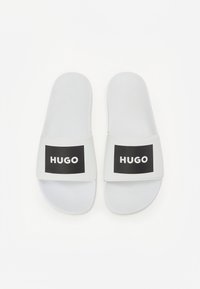 Bijele natikače sa ravnim dizajnom, teksturiranim đonovima i crnim pravokutnim oznakama s natpisom "HUGO" u bijeloj boji.