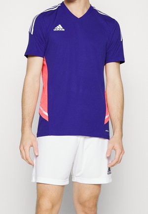 Mężczyzna ubrany w niebieską sportową koszulkę Adidas z białymi paskami na ramionach oraz białe spodenki Adidas.