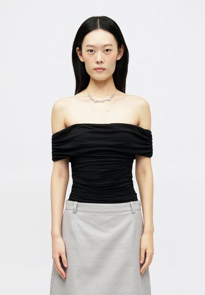 Femme avec des cheveux noirs lisses portant un haut noir à volants dénudés, une jupe grise et un collier en diamant floral sur fond blanc uni.