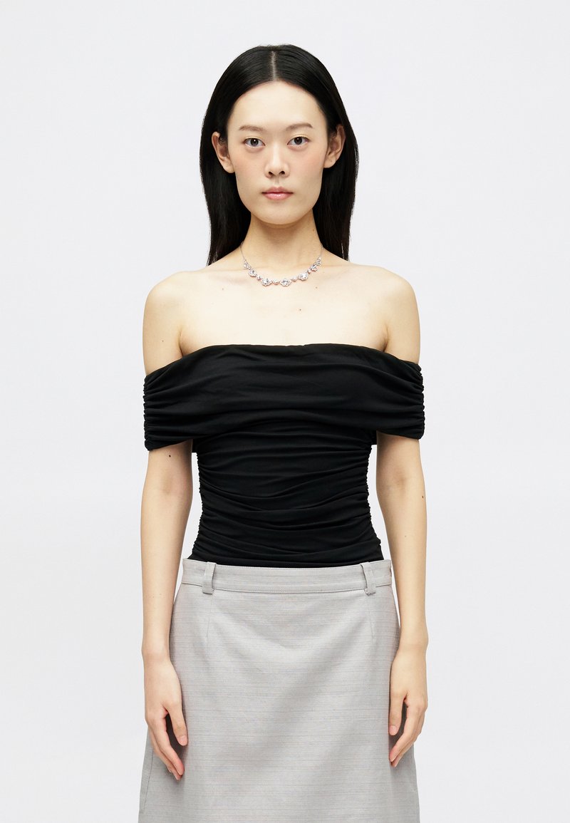 Moteris su tiesiais juodais plaukais, vilkinti raukšlėtą juodą viršų be petnešų, pilkų sijoną ir gėlių formos deimantų vėrinį, prieš baltą fono.