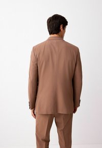 Veste de costume marron en tissu lisse, avec une fente unique au dos, des revers crantés et des accents de boutons contrastants, accompagnée d'un pantalon assorti.
