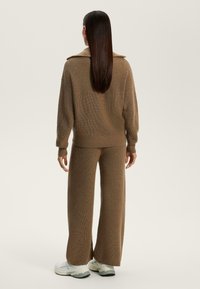 Set de loungewear tricotat, de culoare maro, cu un top lejer cu guler și pantaloni largi. Textura este striată, completată cu adidași ușori.