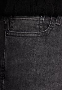 Tmavě šedé džíny z denimu mají přední kapsu s kontrastním prošitím a kovovým knoflíkem, který ukazuje hladkou, texturovanou látku.