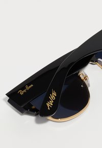 Crne Ray-Ban sunčane naočale s zlatnim detaljima, tamnim lećama i sjajnim završetkom. Imaju odvažni potpis na kraku i izdržljiv dizajn šarke.
