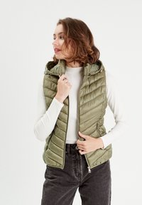 Gilet matelassé vert olive avec des coutures horizontales, une capuche et des poches zippées, porté sur un haut à manches longues à rayures blanches et un jean foncé.