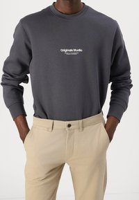 Grå sweatshirt med rund halsringning och "Originals Studio"-logotyp, kombinerad med beige byxor, som visar en avslappnad passform och mjuk tygtextur.