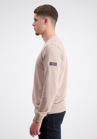 Felpa beige chiaro con collo rotondo, maniche lunghe, polsini e orlo a costine, dotata di una toppa logo blu scuro sul braccio sinistro.