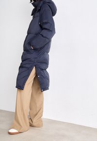 Gepolsterter, kapuzengeführter Mantel in Marineblau mit langer Länge, seitlichen Schlitzen und Druckknopfverschlüssen. Getragen über hellbeigen Hosen und Sandalen.