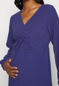 Fialové, strukturované těhotenské šaty s výstřihem do V, dlouhými rukávy a detailem na zavázání v pase, navržené pro pohodlí a pohodlné nošení během těhotenství.