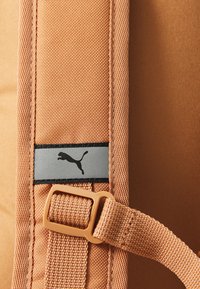 Oranžs mugursomas audums ar strukturētu apdari, kurā ir melna un sudraba Puma logo etiķete un beižs regulējams siksnas sprādze.