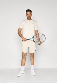 Light beige athletic shirt and shorts, tennis racket in hand, textured fabric, short sleeves, navy logo detail, white sneakers, standing pose.