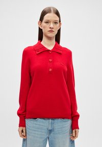 Červený pletený sveter so širokým golierom, zapínaním na štyri gombíky, rebrovanými manžetami a spodným lemom, nosený cez svetlomodré džínsy. Hladká textúra s detailom loga.