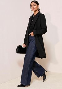 Palton lung negru peste un top texturat negru, asortat cu blugi evazați de un albastru închis și pantofi cu vârf ascuțit negri. Accesorii cu o geantă neagră strălucitoare.