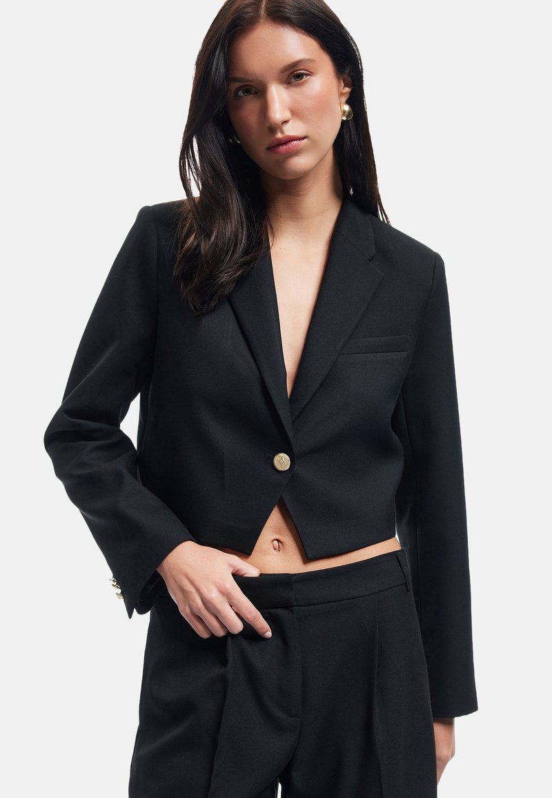 Kort zwart blazer met een enkele gouden knop, notch revers en een zak; gecombineerd met op maat gemaakte zwarte broeken, die een zachte stoftextuur laten zien.