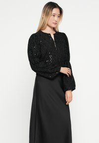 Črna, teksturirana bluzica z napihnjenimi rokavi in sequinskimi poudarki, v kombinaciji s preprosto, dolgimi črnimi krilom; preprost ovratnik in oprijet silhuet.