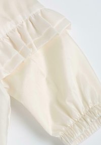 Tissu couleur crème avec des accents froncés, des poignets élastiqués et une texture lisse, idéal pour des vêtements doux et légers.