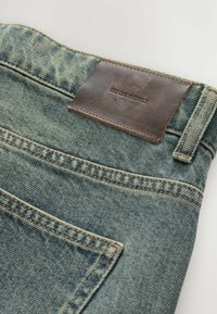 Jean en denim avec un délavage bleu, présentant des coutures contrastantes, une étiquette en cuir portant la mention "DENIM SUPPLY", et deux poches arrière.