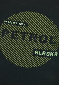 Schwarzes T-Shirt mit einem kreisförmigen grünen grafischen Motiv mit diagonalen Streifen. Der Text enthält "PETROL", "WRECKING CREW" und "ALASKA" in fett gedruckten Schriften.