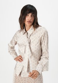 Chemise à manches longues en tissu crème clair avec des rayures verticales et un motif marron répété, dotée d'un col pointu et d'un détail de cravate.