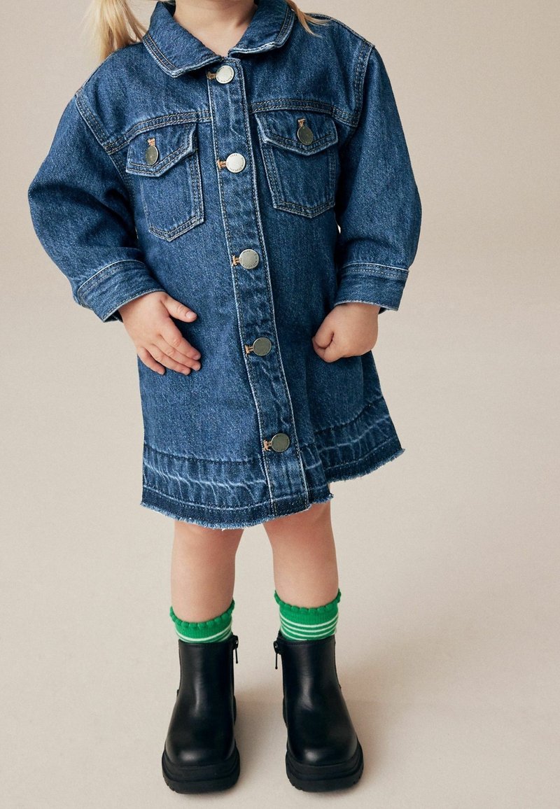 Enfant portant une robe boutonnée en denim bleu, des chaussettes rayées vertes et des bottines noires, debout sur un fond neutre.