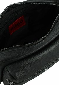 Zwarte leren tas met een textuur, voorzien van een rits en een rode "HUGO" label, met een gladde, glanzende voering.