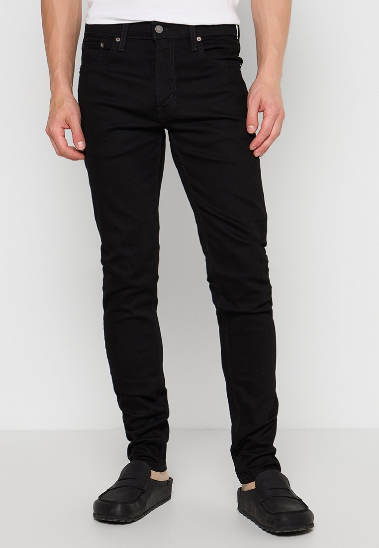 Jeans neri slim-fit realizzati in denim, dotati di tasche frontali, chiusura con bottone e dettagli di cucitura discreti. Indossati con scarpe nere slip-on.