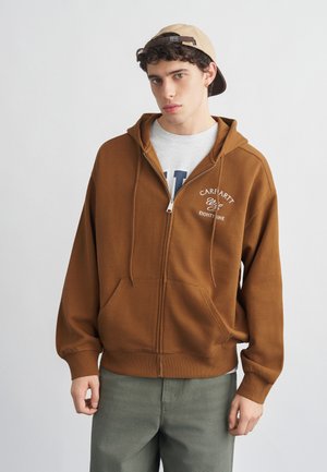 Νεαρός άνδρας που φοράει μπεζ καπέλο με το γείσο προς τα πίσω, καφέ φούτερ με φερμουάρ και το λογότυπο "Carhartt", ανοιχτό γκρι μπλουζάκι και ελιά παντελόνι, στέκεται μπροστά σε απλό φόντο.