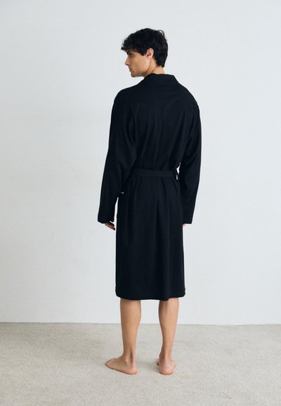 Robe noire en tissu doux, de longueur mi-mollet avec de longues manches, une ceinture à la taille et un col au design simple.